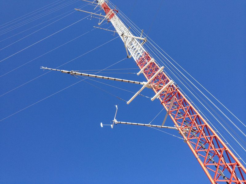 A guyed met mast carrying anemometers and wind vanes on booms, against a clear blue sky